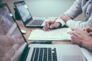 Close up image of two people working side by side on their laptops reviewing notes between them